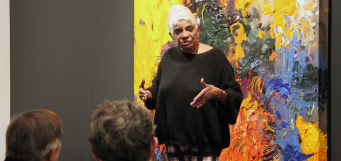 A medium-skinned woman with bright white hair stands in front of a colorful abstract painting speaking to a crowd of visitors.