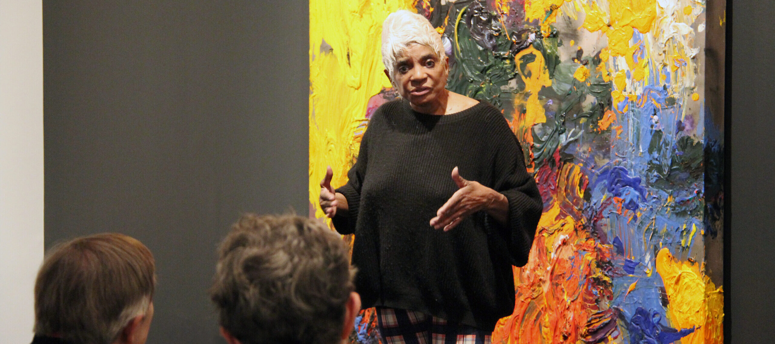 A woman with a medium-dark skin tone and white, short hair, is standing in front of a colorful abstract painting talking to an audience.