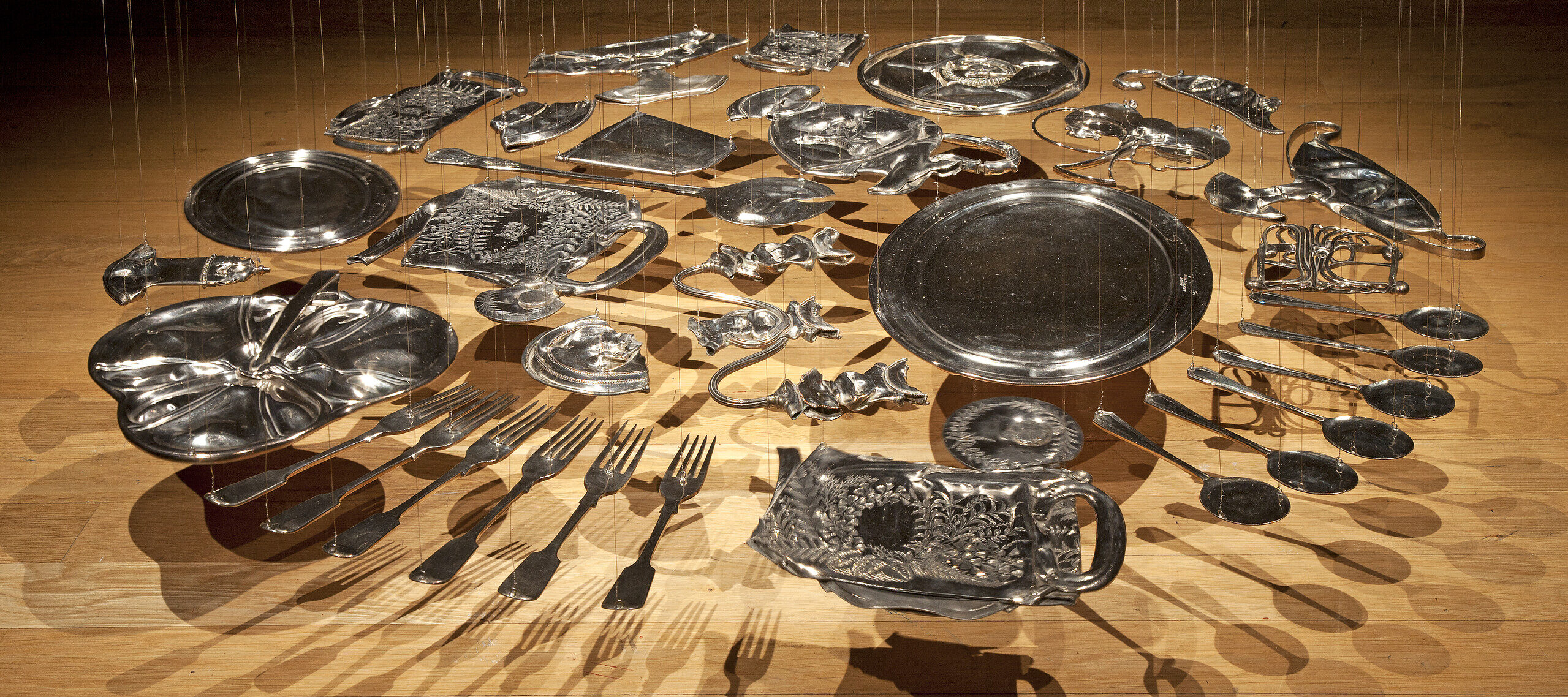 Thirty silver-plated items, including spoons, forks, trays, and various serving ware, are flattened and hang suspended from the ceiling with thin, nearly invisible wire. The pieces are arranged in a circle several inches above the floor.