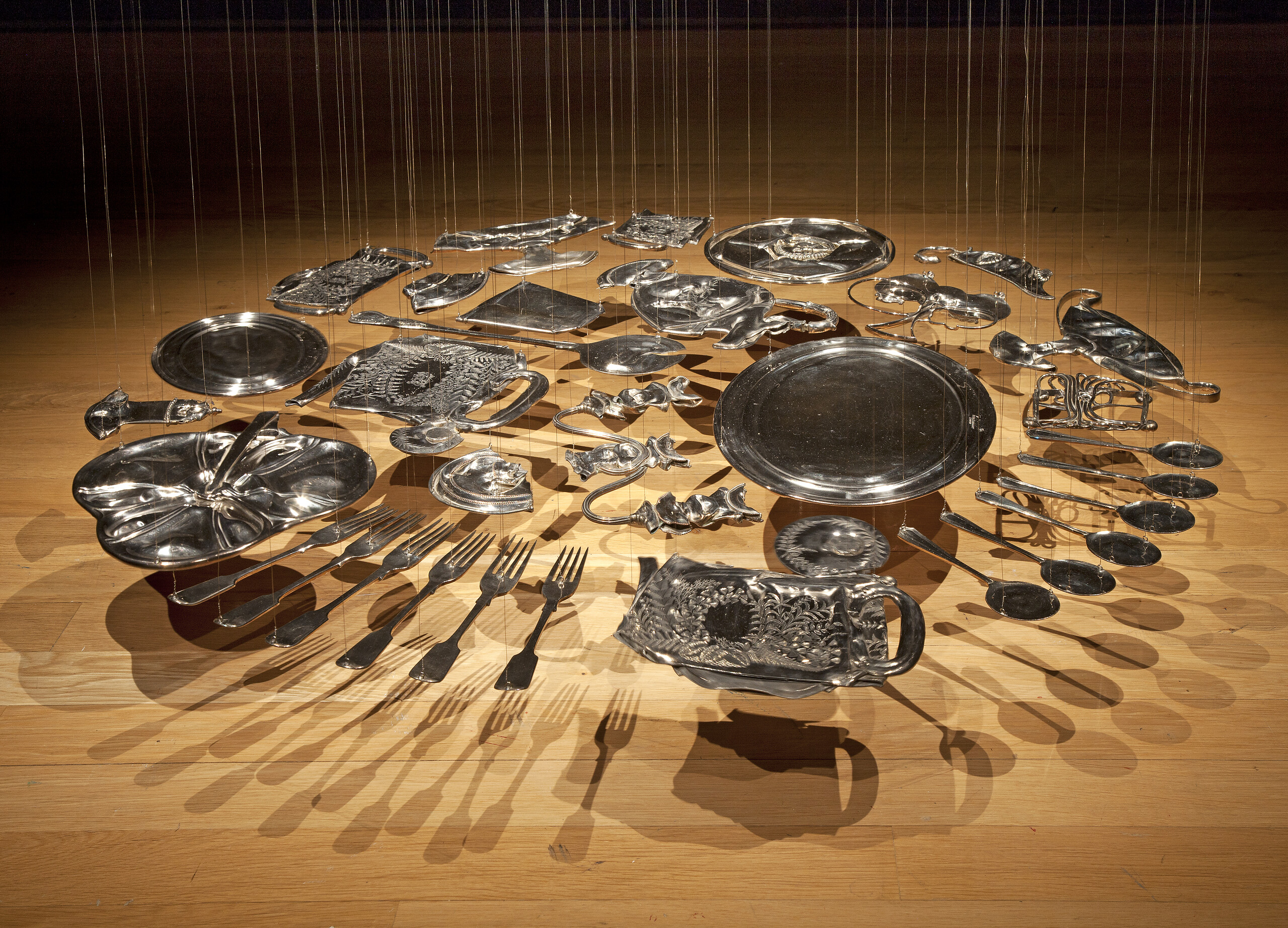 Thirty silver-plated items, including spoons, forks, trays, and various serving ware, are flattened and hang suspended from the ceiling with thin, nearly invisible wire. The pieces are arranged in a circle several inches above the floor.