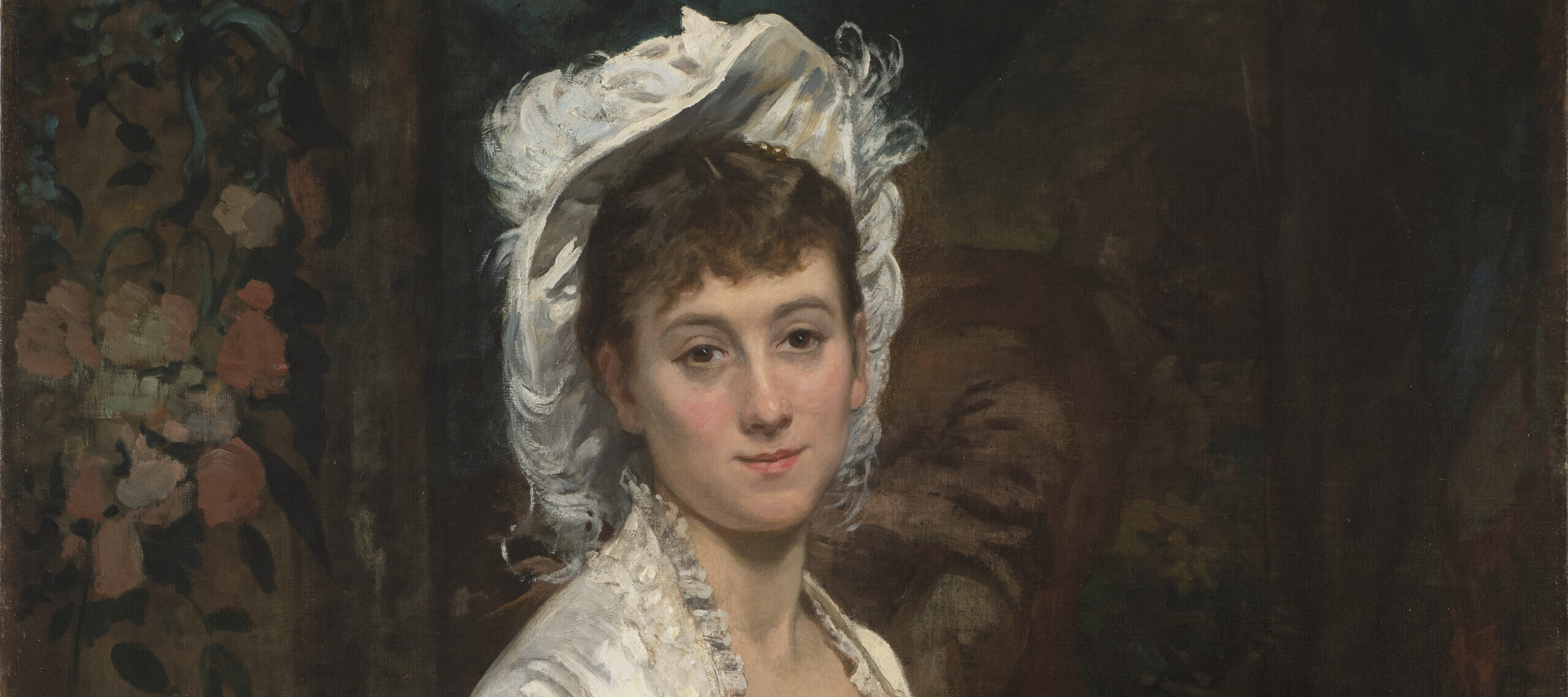 A woman with light skin tone and brown hair sits and looks at the viewer with a slight smile and calm expression, her hands resting in her lap. In a painting with loose brushstrokes, her face is more realistically detailed than her dress and darker interior surroundings. She wears an old-fashioned, ornate white silk gown with ruffles at the bodice and skirt, as well as a matching, upturned white hat with feathers and frills.