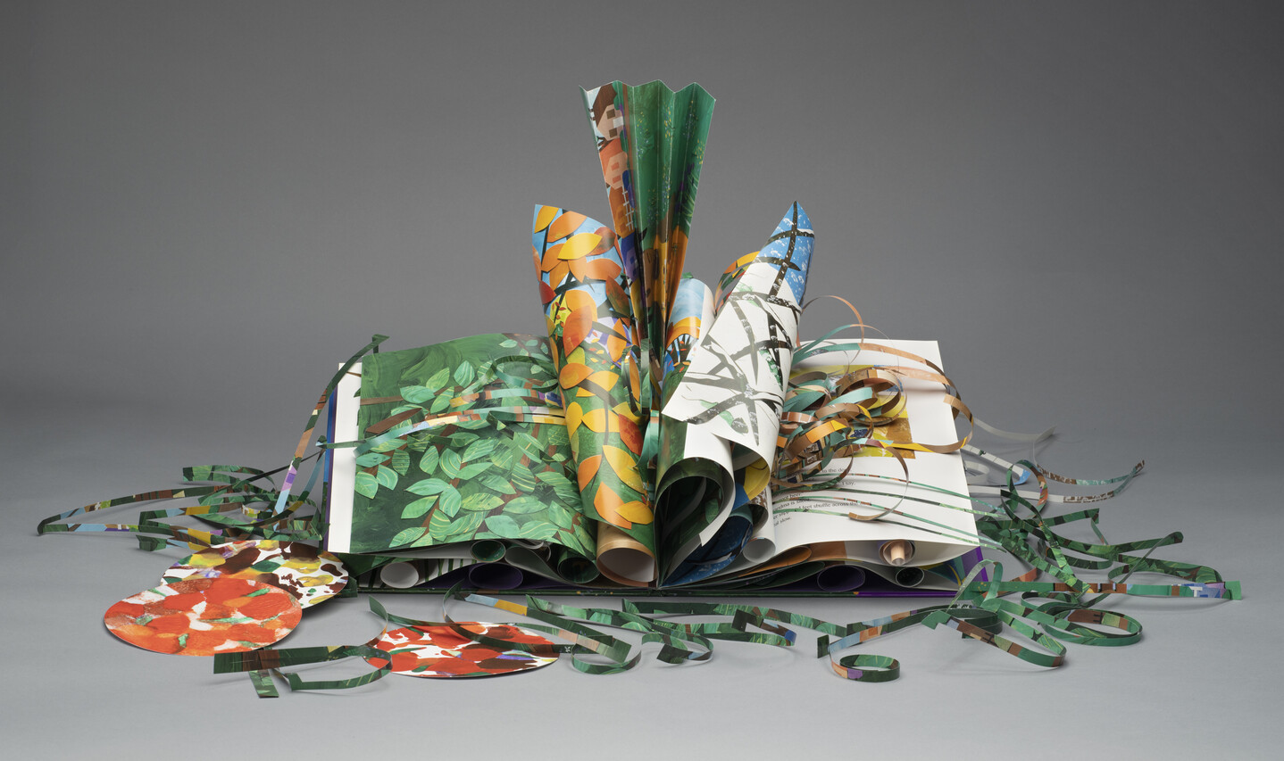 An artist's book made of colorful green, yellow, brown, orange, and red paper. Several of the pages are curled and folded. Other paper is cut into strips and circles and displayed around the book.