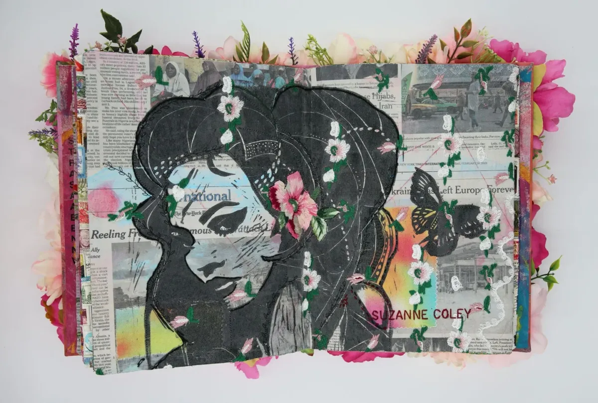 A photo of an artist's book, open in the middle. The pages are made from newspaper with embroidered white and pink flowers and an image of a woman's face and butterfly printed in black ink.