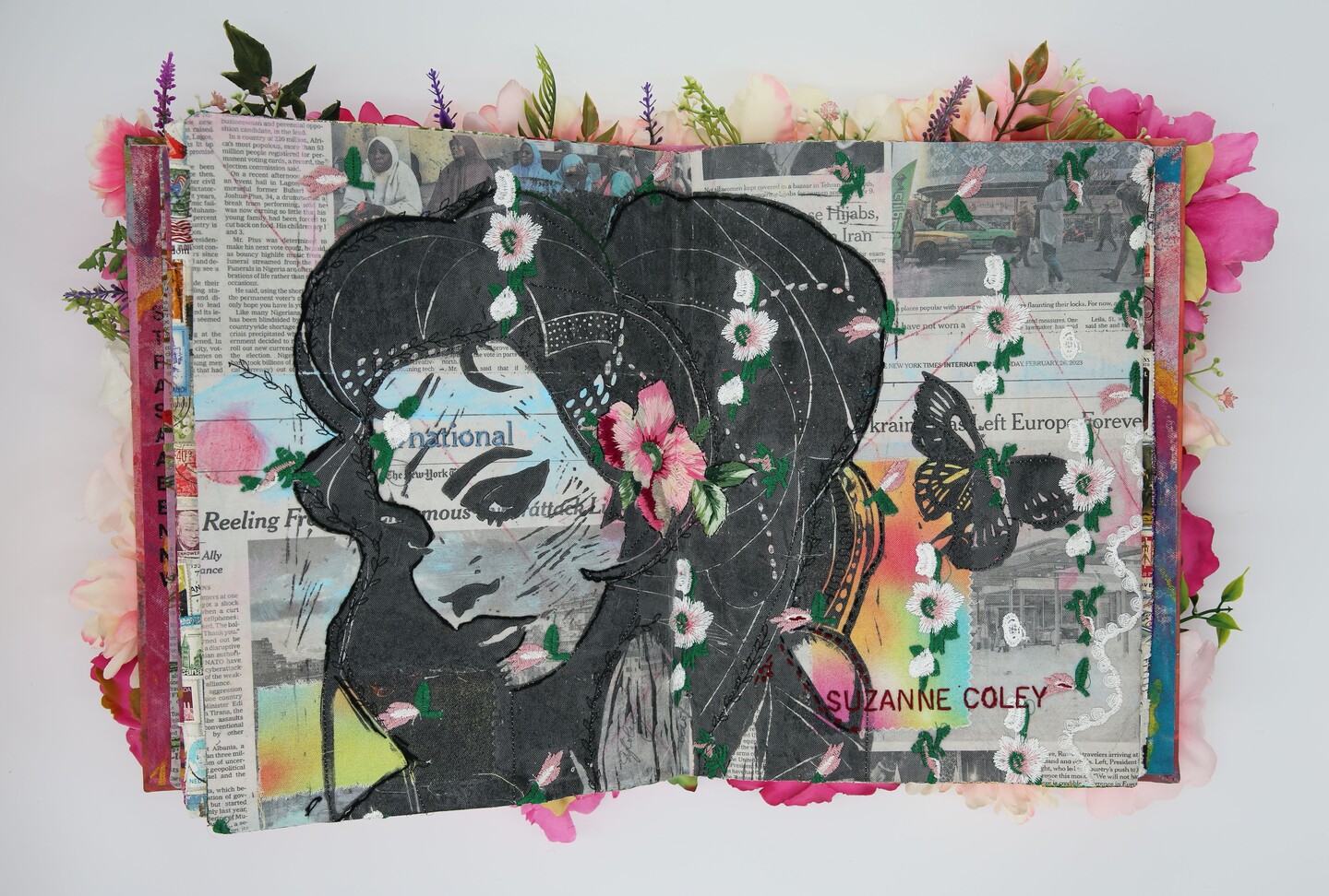 A photo of an artist's book, open in the middle. The pages are made from newspaper with embroidered white and pink flowers and an image of a woman's face and butterfly printed in black ink.