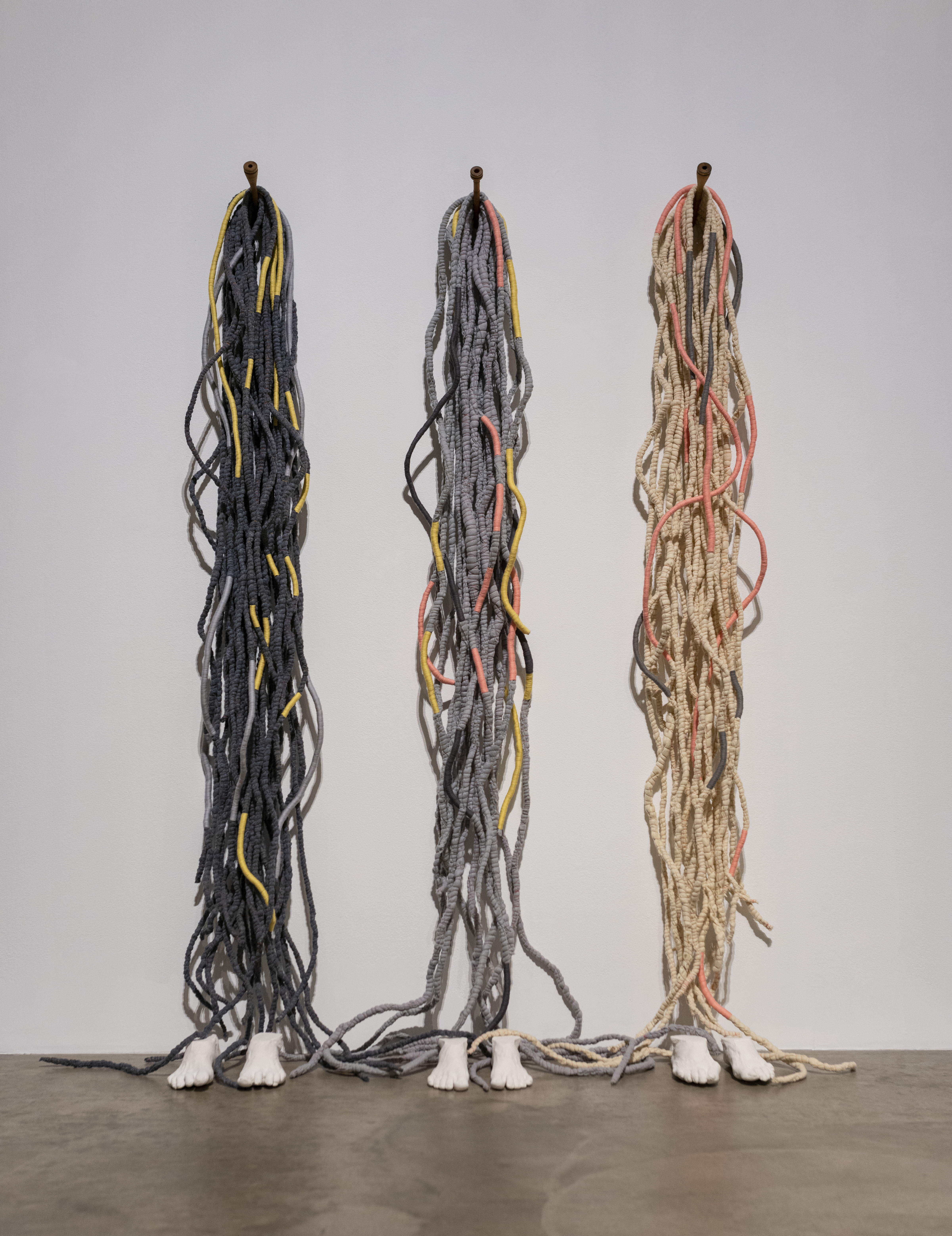 Three sets of different colored rope – beige, gray, and black – are equidistantly nailed to a wall. On the floor at the bottom of each set of rope is a pair of individualized white plaster cast feet.