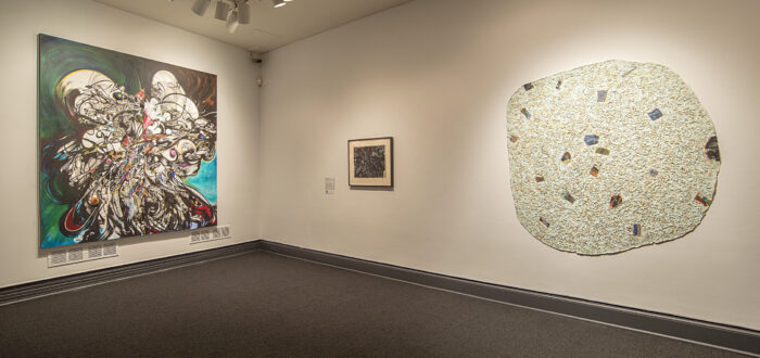 An installation photograph of a two artworks hanging next to each other on a white wall. The artwork on the right is a large round-shaped abstract form that has an organic quality. It is white, with speckles, and has a rough texture to it, evoking the texture of sand.
