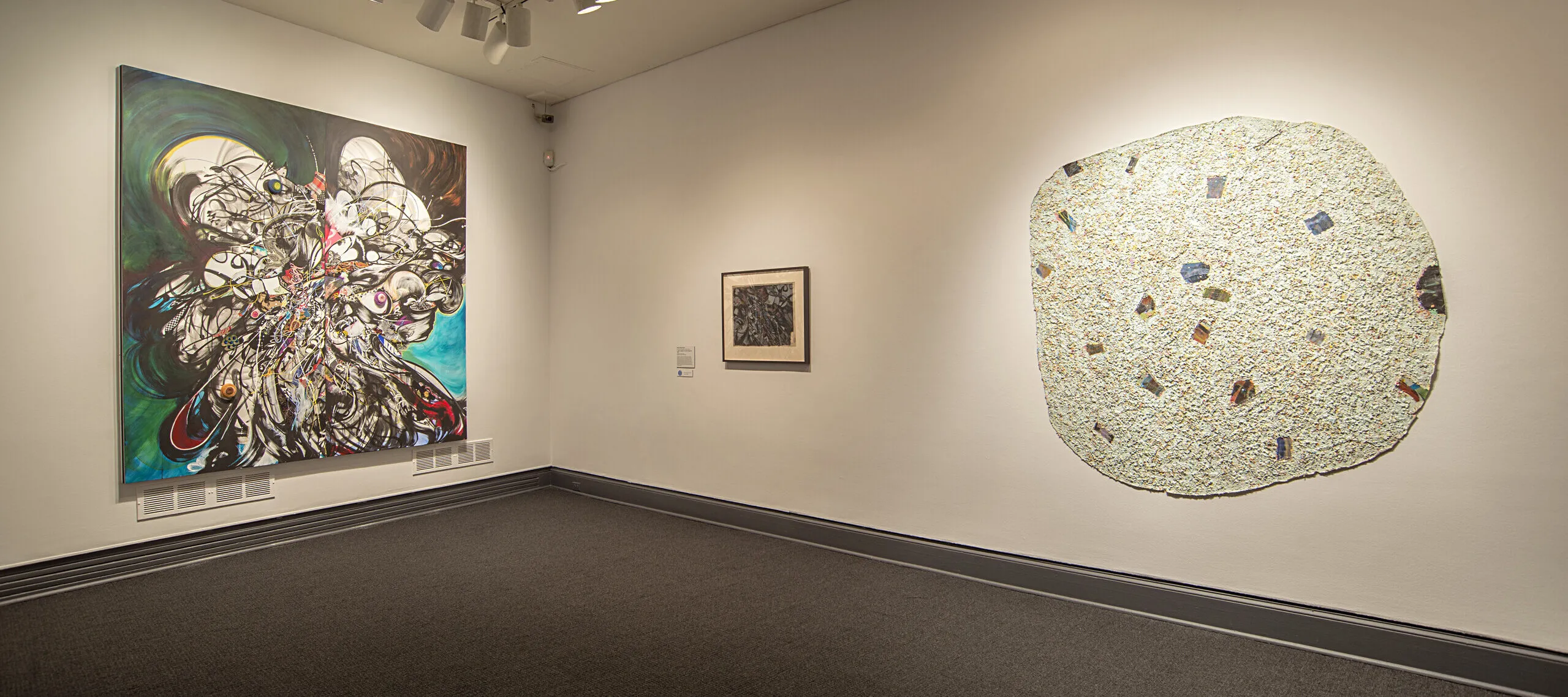 An installation photograph of a two artworks hanging next to each other on a white wall. The artwork on the right is a large round-shaped abstract form that has an organic quality. It is white, with speckles, and has a rough texture to it, evoking the texture of sand.