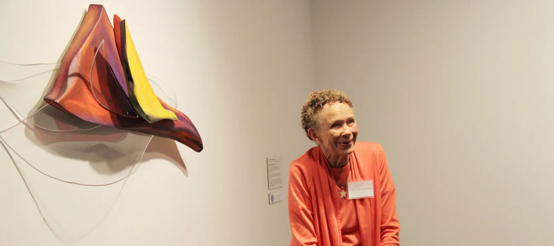 A woman with medium-dark skin is standing before an artwork hanging on a white wall. The artwork is a triangular, organic, sculptural piece in red and yellow tones.