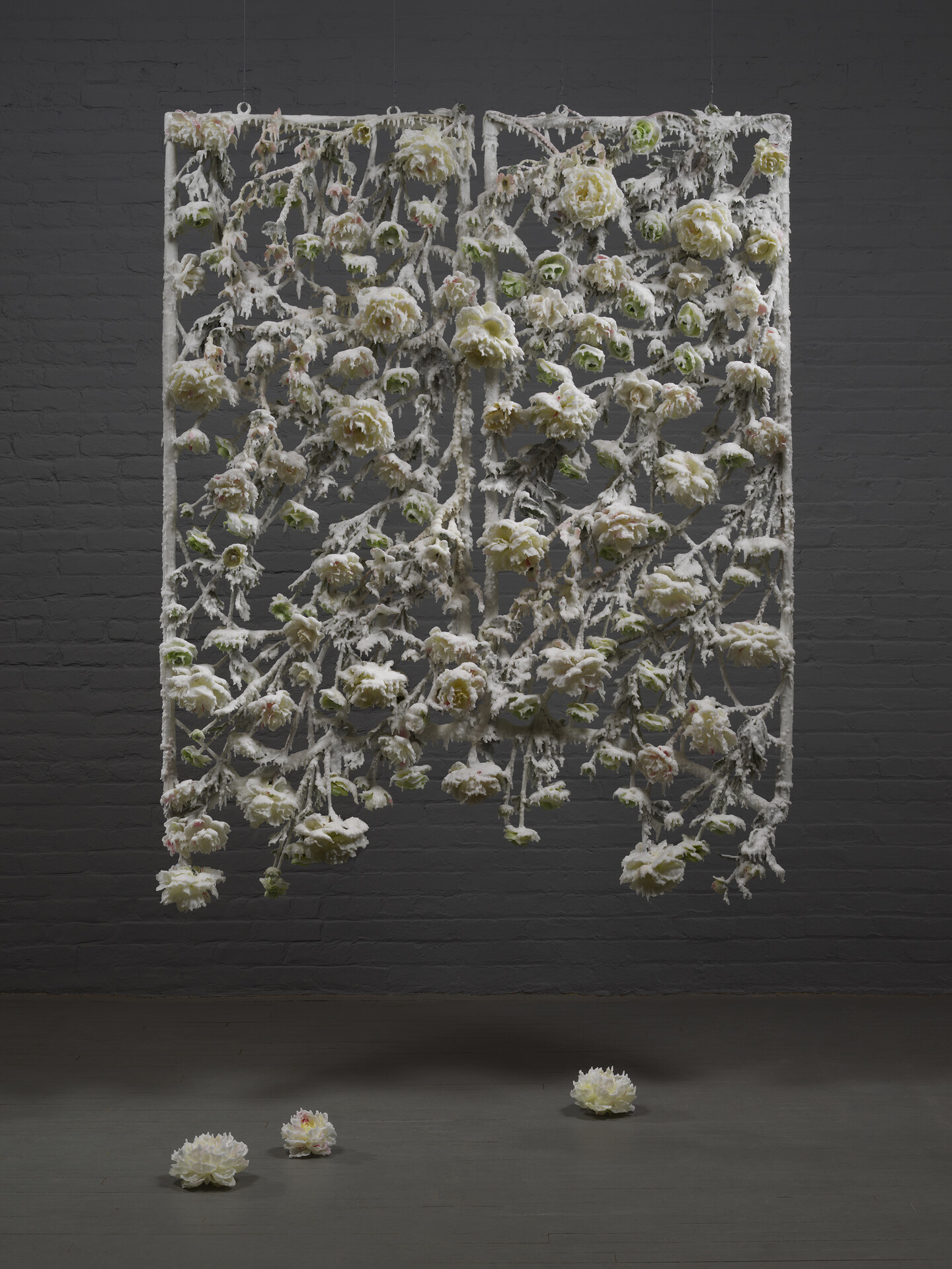 A sculpture suspended from the ceiling made of wax-covered silk flowers hanging within a rectangular steel frame. Hardened white wax coats and drips down all surfaces of the sculpture against a dark grey background.