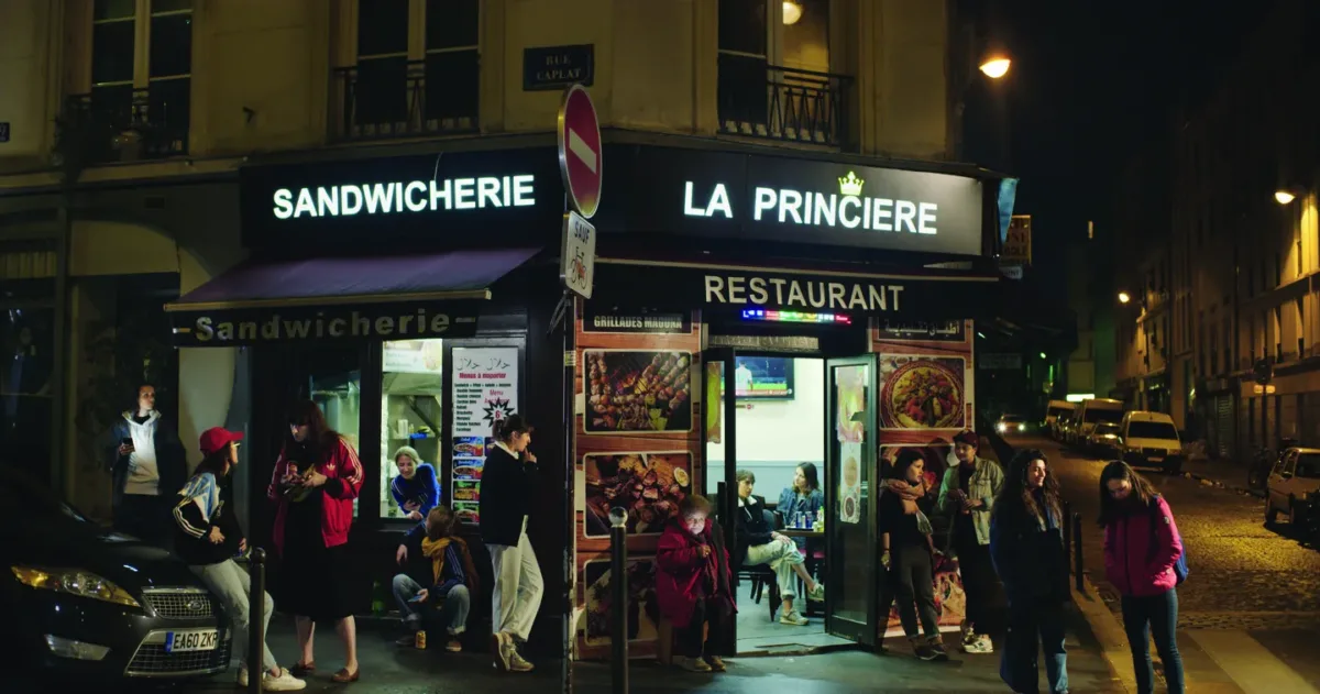 Group of women gathered outside of a shop on the corner at night. The lighted signs above the shop read “Sandwichiere” and “La Princiere”