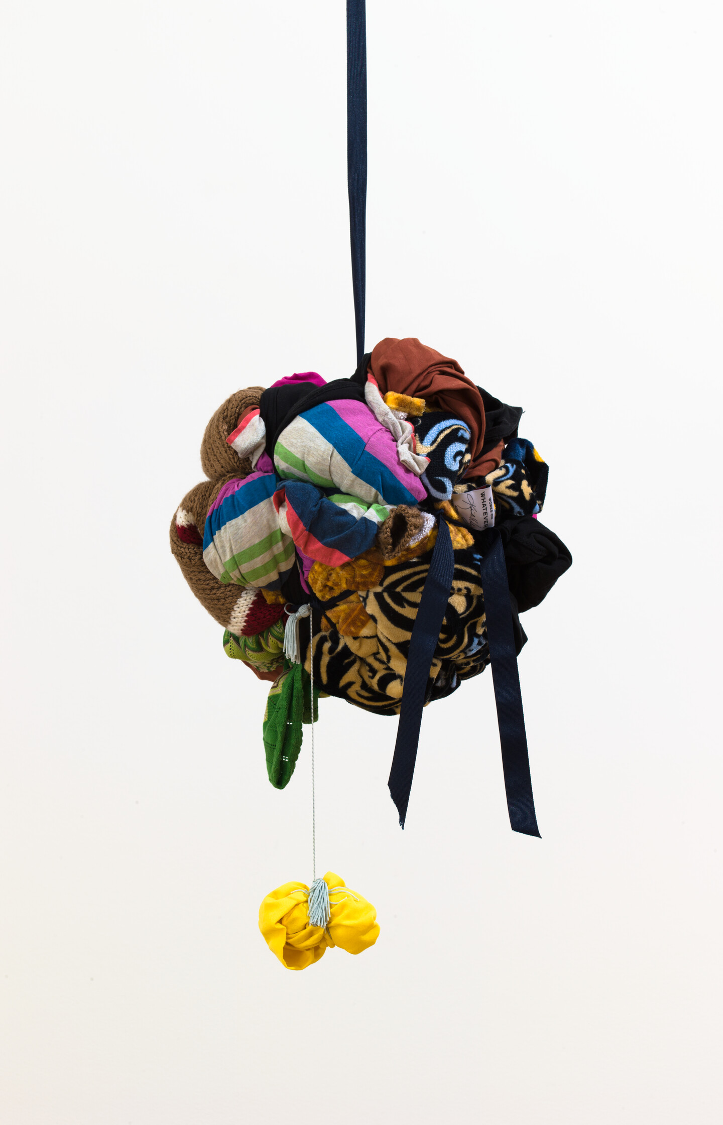 A sculpture made of colorful fabric tied into a round form hanging from a strip of black fabric. A smaller ball of yellow fabric hangs below with a ribbon.