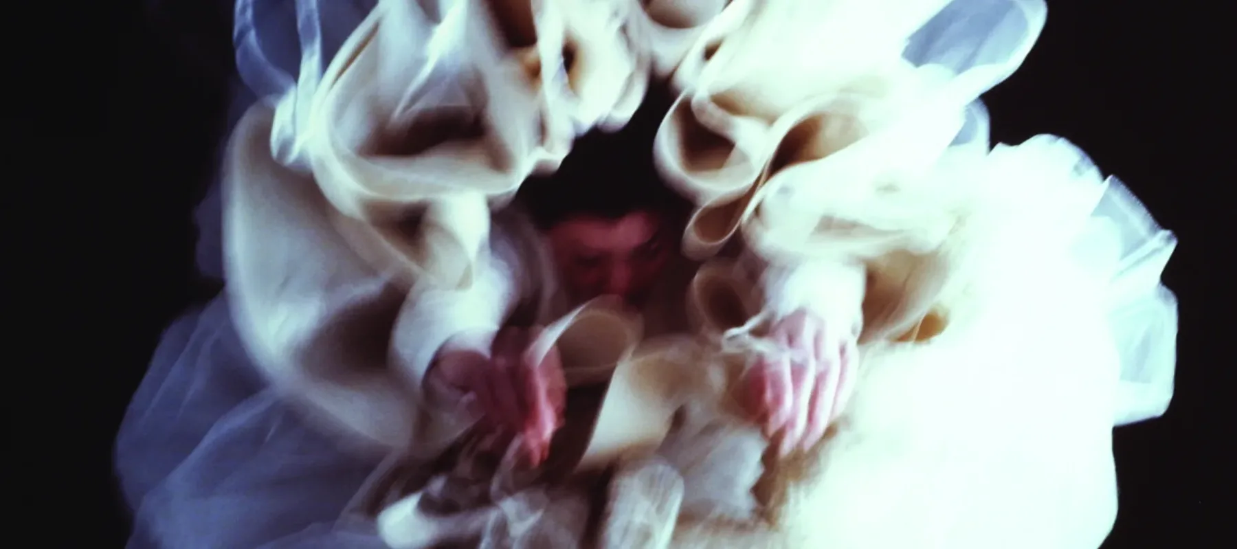 Large, glossy, blurry, color photograph of a light-skinned woman hanging upside-down directly above the viewer.  Her arms hang loosely above her head, enveloped in diaphanous white skirts billowing around her, set against a black background, creating a flower-like appearance.