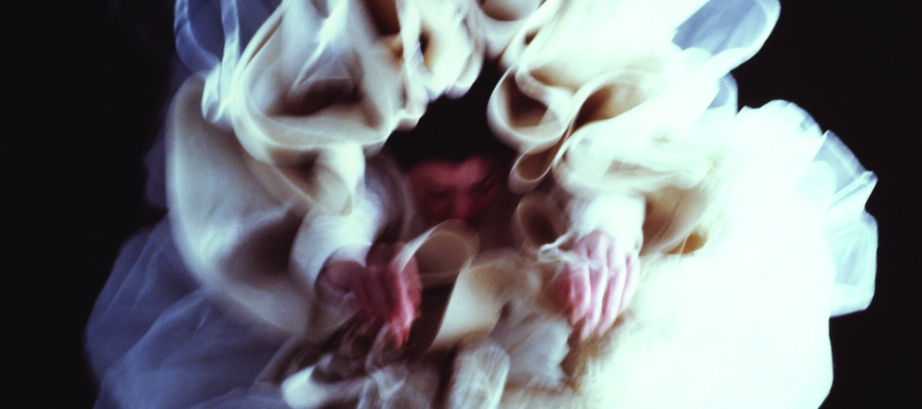 Large, glossy, blurry, color photograph of a light-skinned woman hanging upside-down directly above the viewer.  Her arms hang loosely above her head, enveloped in diaphanous white skirts billowing around her, set against a black background, creating a flower-like appearance.