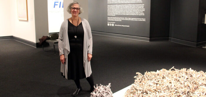 A woman with a light skin tone and gray, short hair is standing in a gallery space next to a sculpture lying on the ground. The sculpture is a large, round shape, and a smaller, round ball of newspapers folded into thin tubes and tied together. The many tubes tied together look like brushes made from newspaper. They are black and white with some pops of color in between.