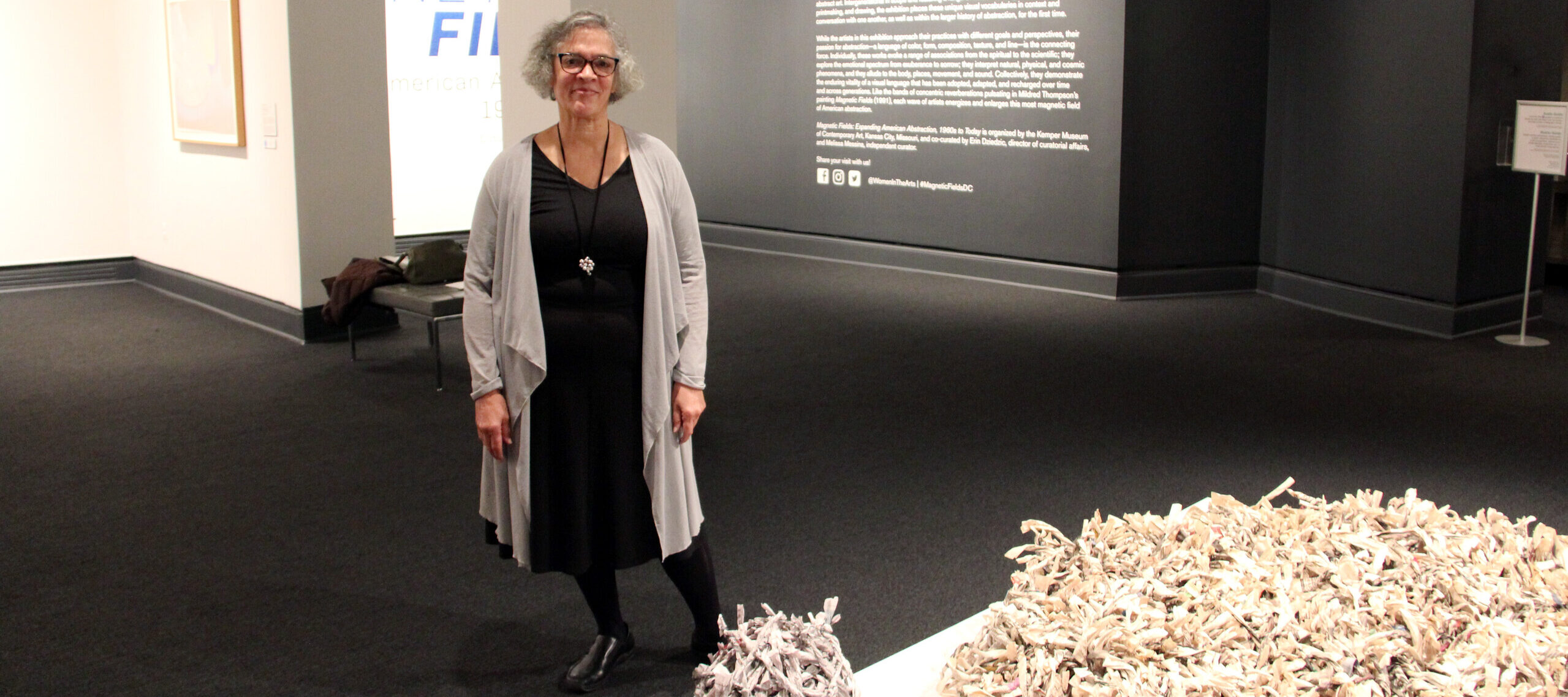 A woman with a light skin tone and gray, short hair is standing in a gallery space next to a sculpture lying on the ground. The sculpture is a large, round shape, and a smaller, round ball of newspapers folded into thin tubes and tied together. The many tubes tied together look like brushes made from newspaper. They are black and white with some pops of color in between.