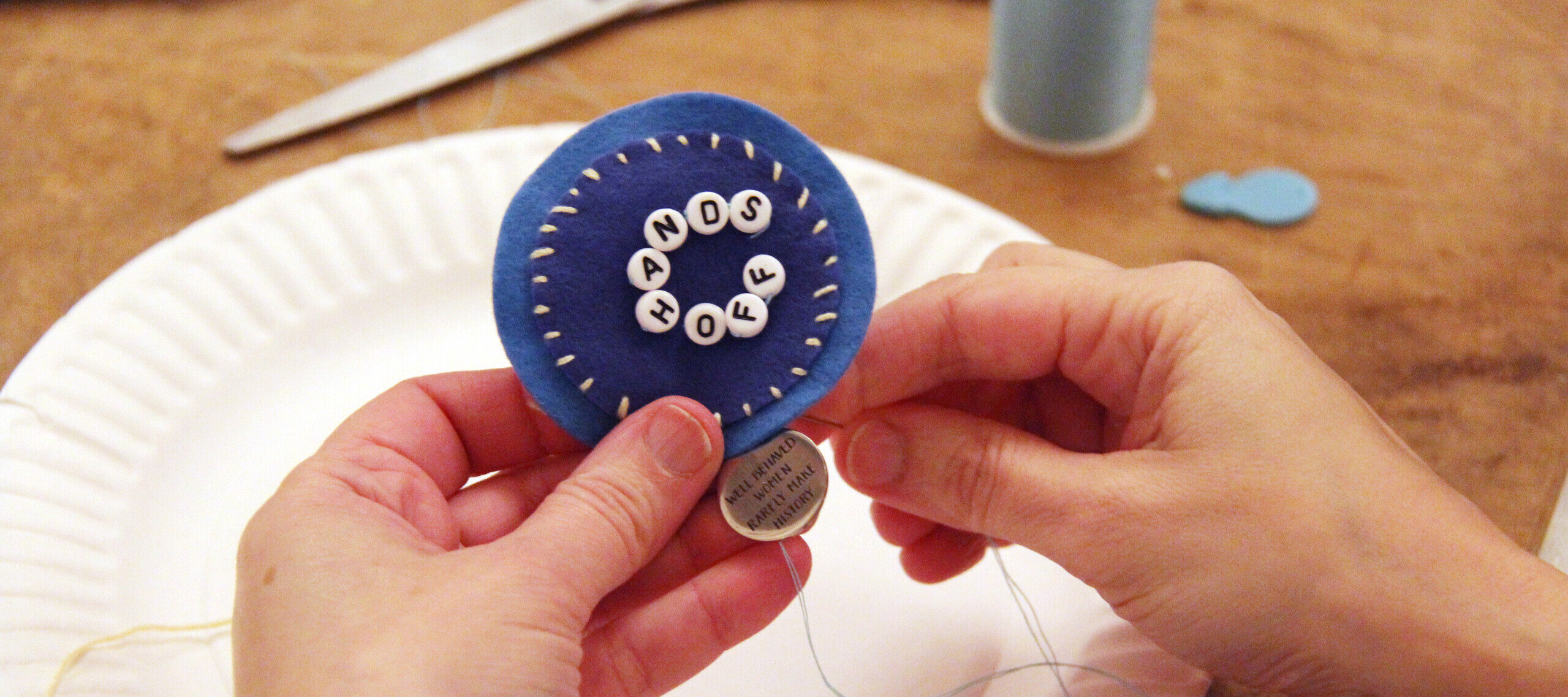 Close-up of a person's hands with light skin tone sewing a pin that reads “hands off.” In the background are scissors, a needle threader, and a spool of light blue thread.