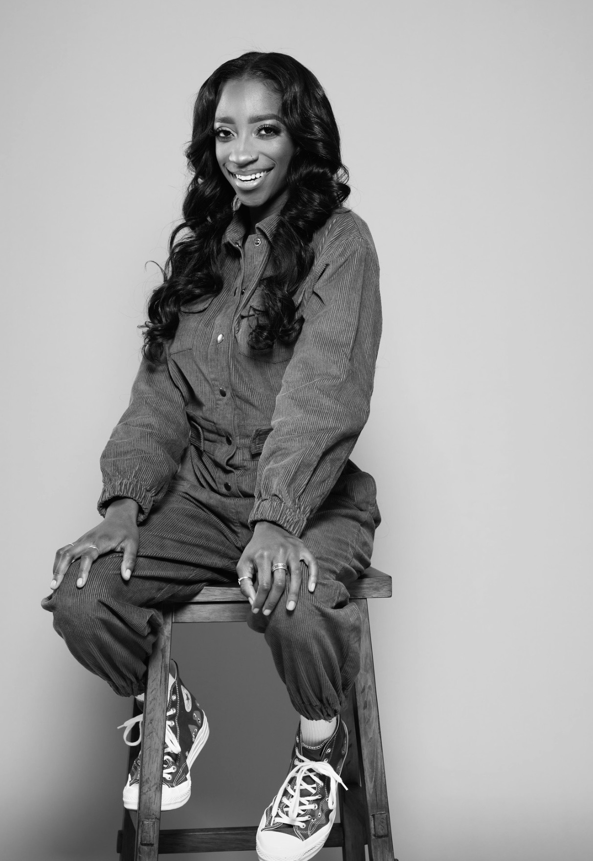 A black-and-white portrait of a woman with a dark skin tone and long, wavy hair. She is wearing a corduroy jumpsuit and is sitting on a wooden chair. 
