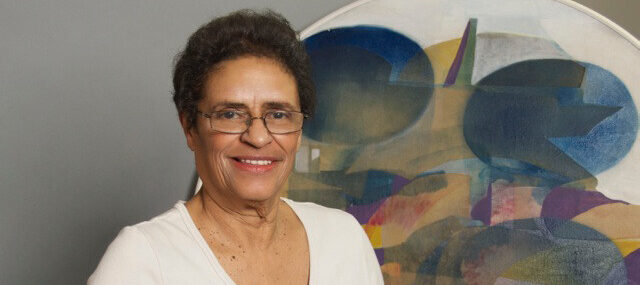 A medium-skinned woman stands before a colorful, abstract painting. She is wearing glasses and a white v-neck sweater, and she is smiling at the camera.
