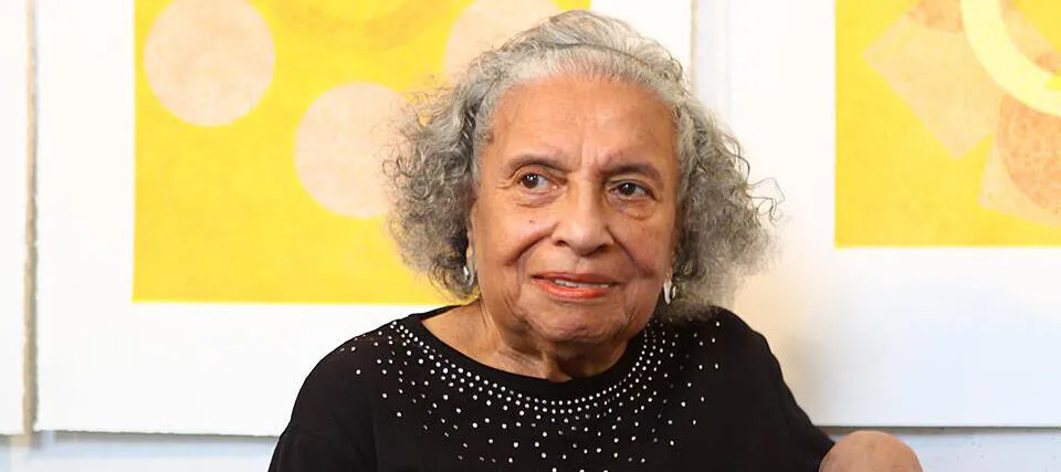 A woman with medium-dark skin and short, gray hair sits before a yellow and white painting. She wears a black shirt with rhinestones and looks slightly to her right, smiling into the distance.