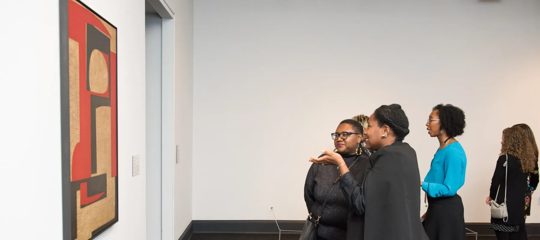 A group of women with a dark medium skin tone are standing in a gallery looking at a large abstract painting and gesturing towards it, discussing the painting. The women look very interested and are having a lively conversation. The painting is a large abstract painting featuring geometric shapes in red, brown, and black.