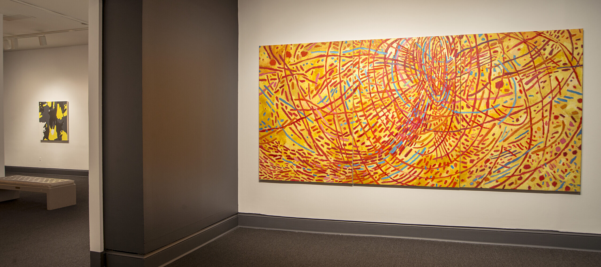 An installation shot of a gallery with a large painting hanging on the wall. Abstract painting features a vivid yellow background covered by circles, daubs, and straight and wavy lines in red, orange, cobalt, sky blue, and violet. Arcing red strokes evoke concentric circles. Straight lines in other hues radiate out from the center circle like a starburst.