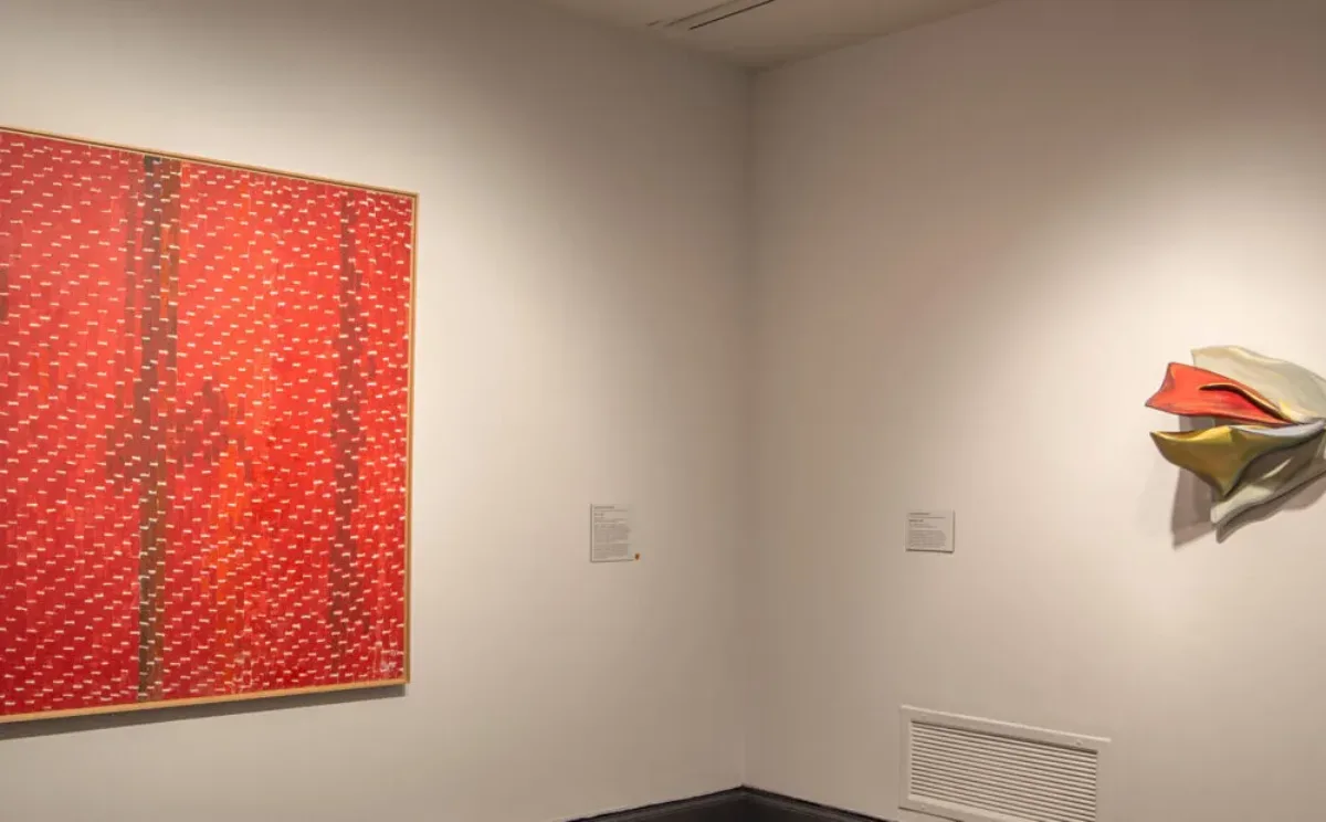 A view of a gallery space with a painting and a sculpture hanging on white walls. The painting on the left is a large, red canvas with little dots in a darker color in vertical lines.