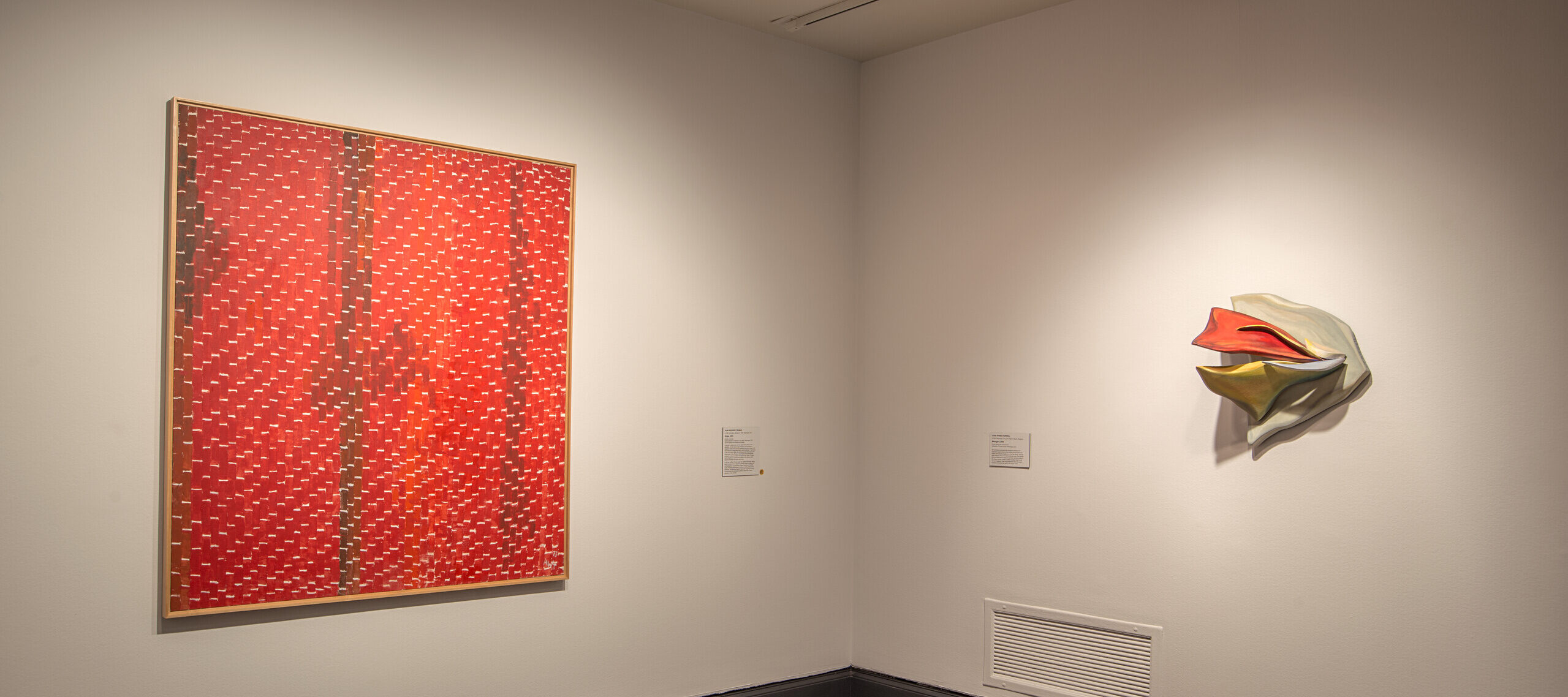 A view of a gallery space with a painting and a sculpture hanging on white walls. The painting on the left is a large, red canvas with little dots in a darker color in vertical lines.