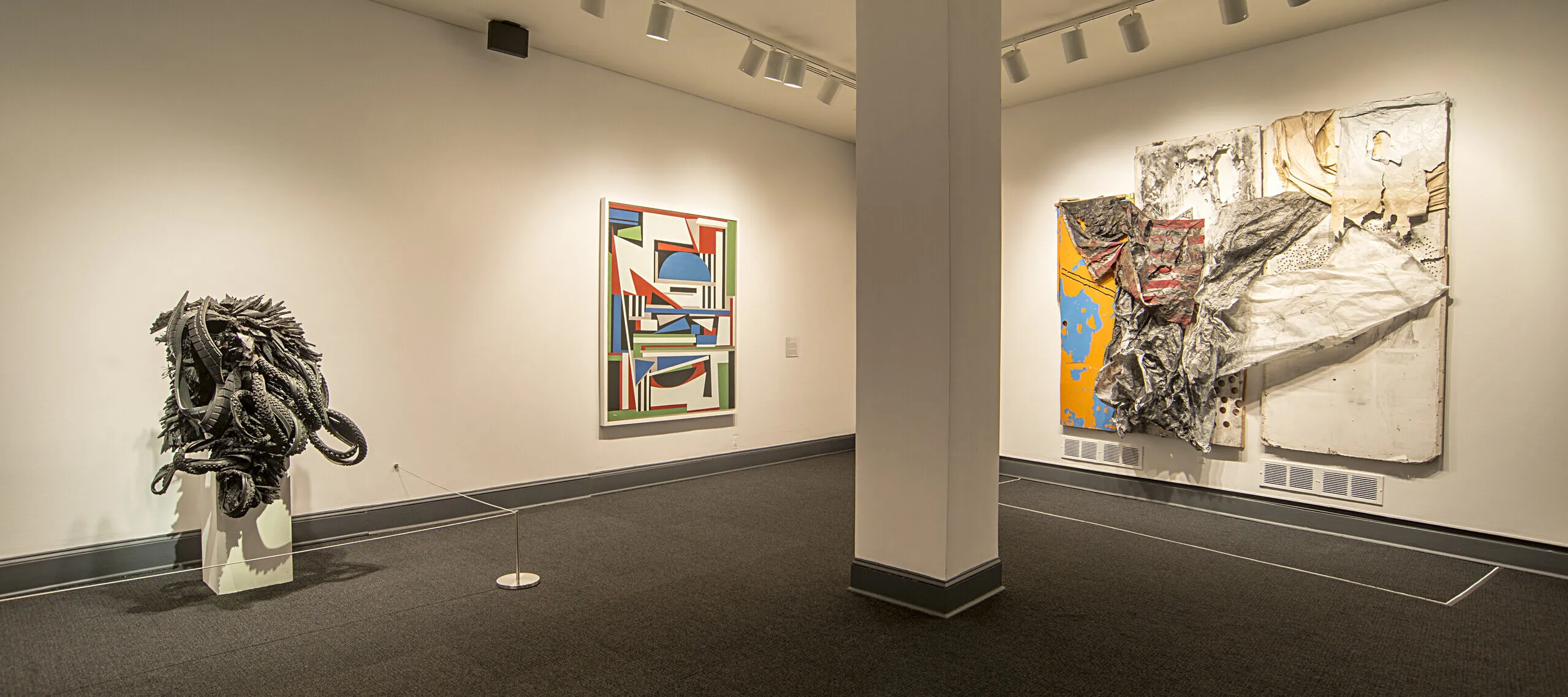 An installation shot of a gallery shows several art pieces in the room. On the left, there is a sculpture made from black rubber tires sits on a. The black rubber tires are shaped and distorted in way that they create an organic form, resembling an insect or an alien-like figure.