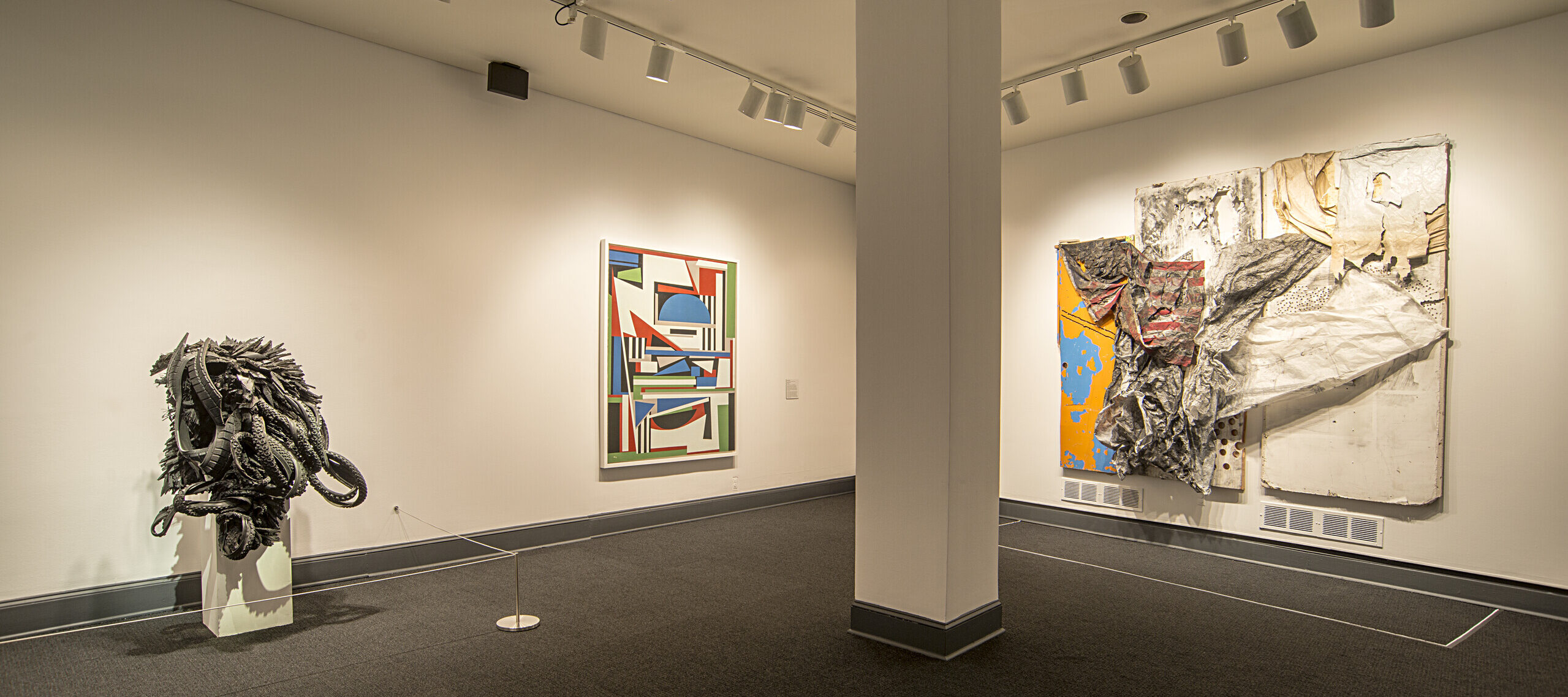 An installation shot of a gallery shows several art pieces in the room. On the left, there is a sculpture made from black rubber tires sits on a. The black rubber tires are shaped and distorted in way that they create an organic form, resembling an insect or an alien-like figure.