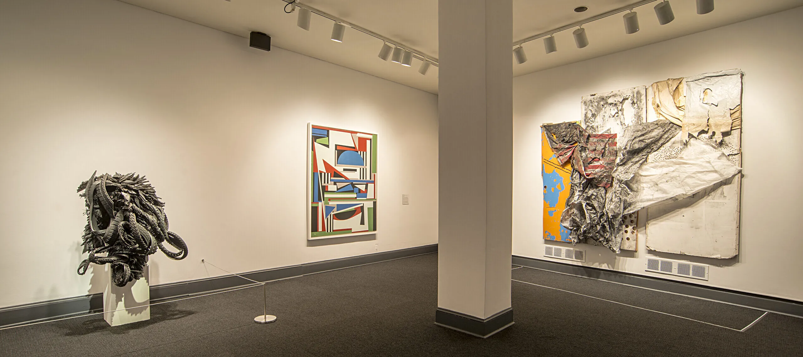 An installation shot of a gallery shows several art pieces in the room. On the left, there is a sculpture made from black rubber tires sits on a. The black rubber tires are shaped and distorted in way that they create an organic form, resembling an insect or an alien-like figure.