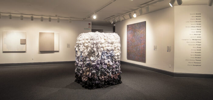 An installation shot of a gallery with a large square sculpture in the middle of the room. The square sculpture is as tall as person and the surface is covered in textiles, possibly shirts, dyed in different colors. On top, the textiles are white, and they slowly fade into a lilac and blue hue. The change of color has an ombre-effect.