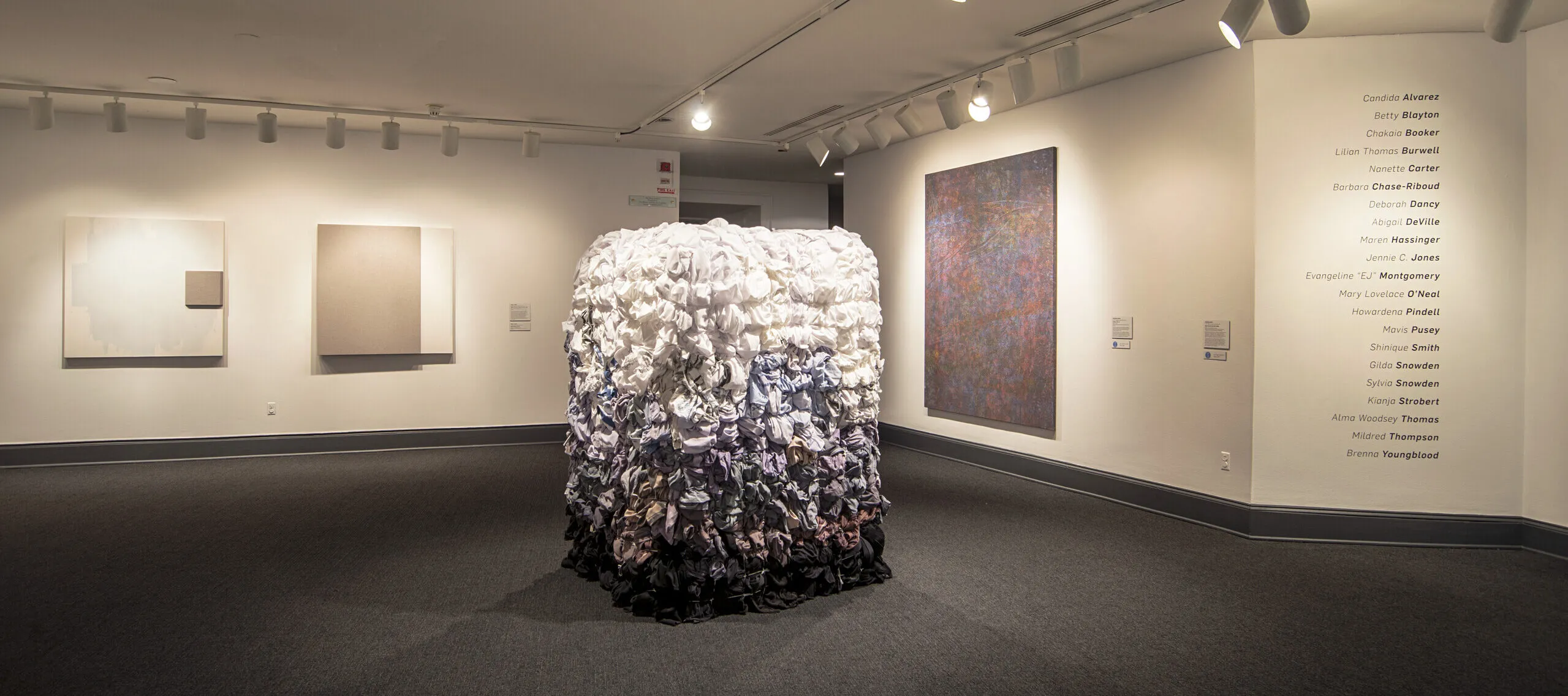 An installation shot of a gallery with a large square sculpture in the middle of the room. The square sculpture is as tall as person and the surface is covered in textiles, possibly shirts, dyed in different colors. On top, the textiles are white, and they slowly fade into a lilac and blue hue. The change of color has an ombre-effect.