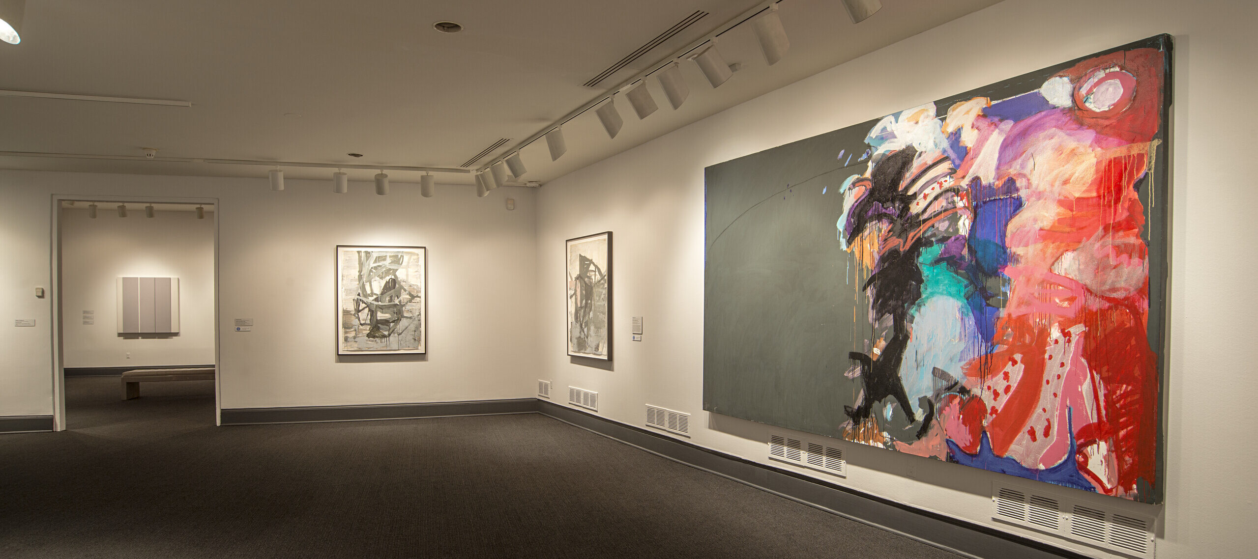 A view of a gallery with white walls and a gray floor. A large canvas with colorful brushstrokes takes up almost the entire wall. On a gray background, brushstrokes in pink, red, blue, and brown take up half of the canvas, while the other half remains gray.