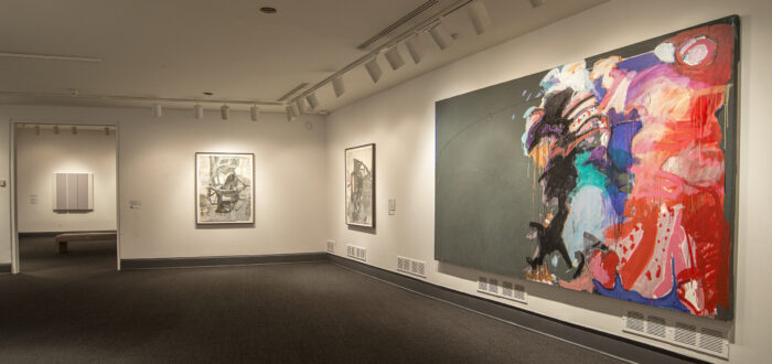 An installation shot of a room with white walls and a large, abstract painting hanging on one of the walls on the right. The large painting is comprised of colorful brushstrokes against a dark gray background.