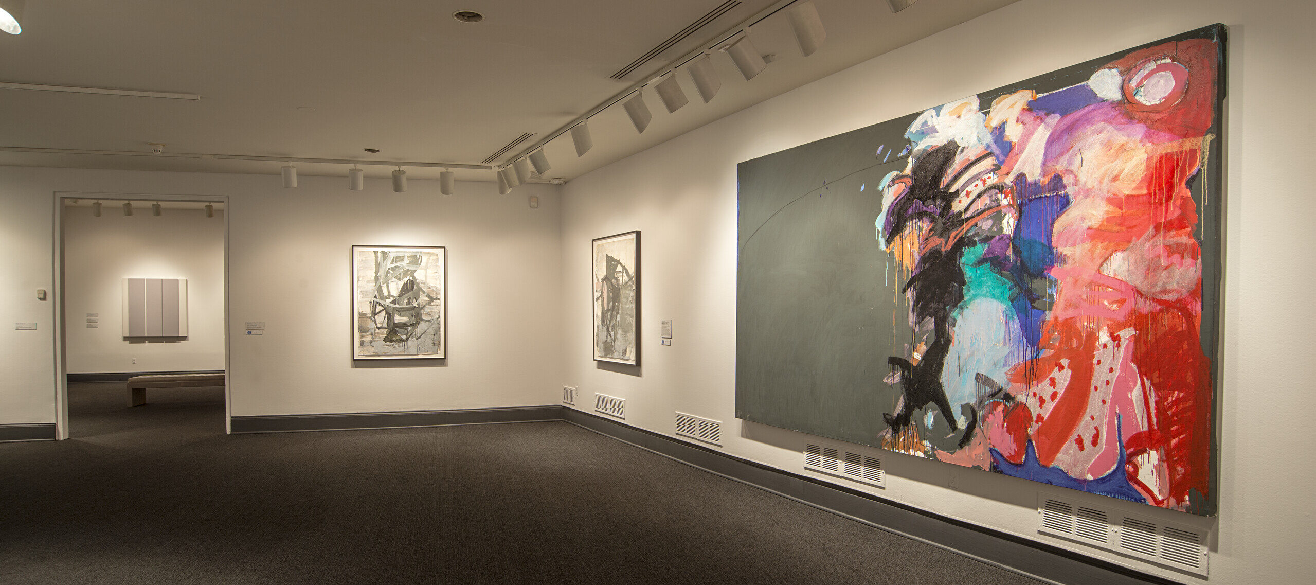 An installation shot of a room with white walls and a large, abstract painting hanging on one of the walls on the right. The large painting is comprised of colorful brushstrokes against a dark gray background.