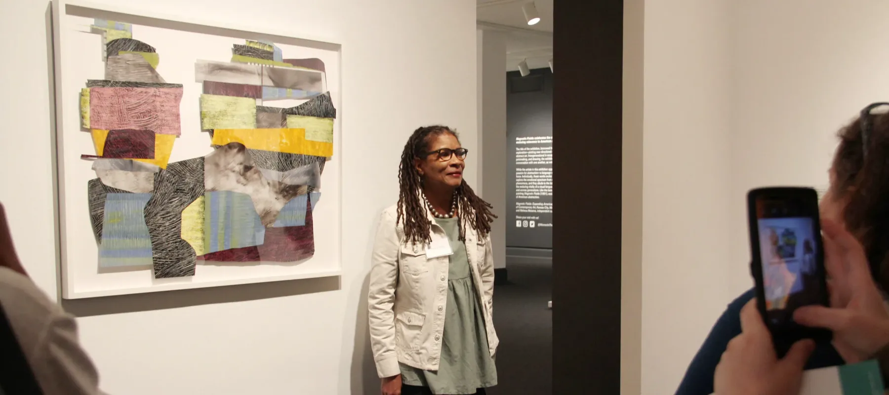 A woman with a medium-dark skin tone and long, dark hair stands in a gallery before a couple of people next to an art piece hanging on the wall. The art piece is a structure made of a variety of shapes stacked on top of each other in different colors and with different textures. The brushstrokes are visible, giving the piece an interesting texture. The colors are vibrant against the white background.