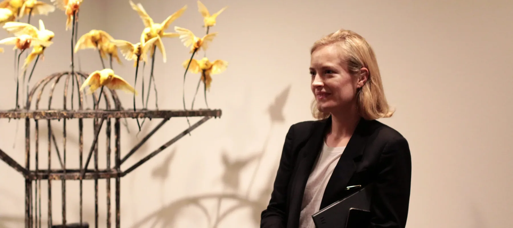 A woman with a light skin tone and short, blonde hair stands before a white wall next to a sculpture. The sculpture is a metal construction with yellow birds attached to its top.