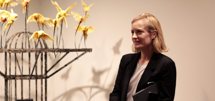 A woman with a light skin tone and short, blonde hair stands before a white wall next to a sculpture. The sculpture is a metal construction with yellow birds attached to its top.