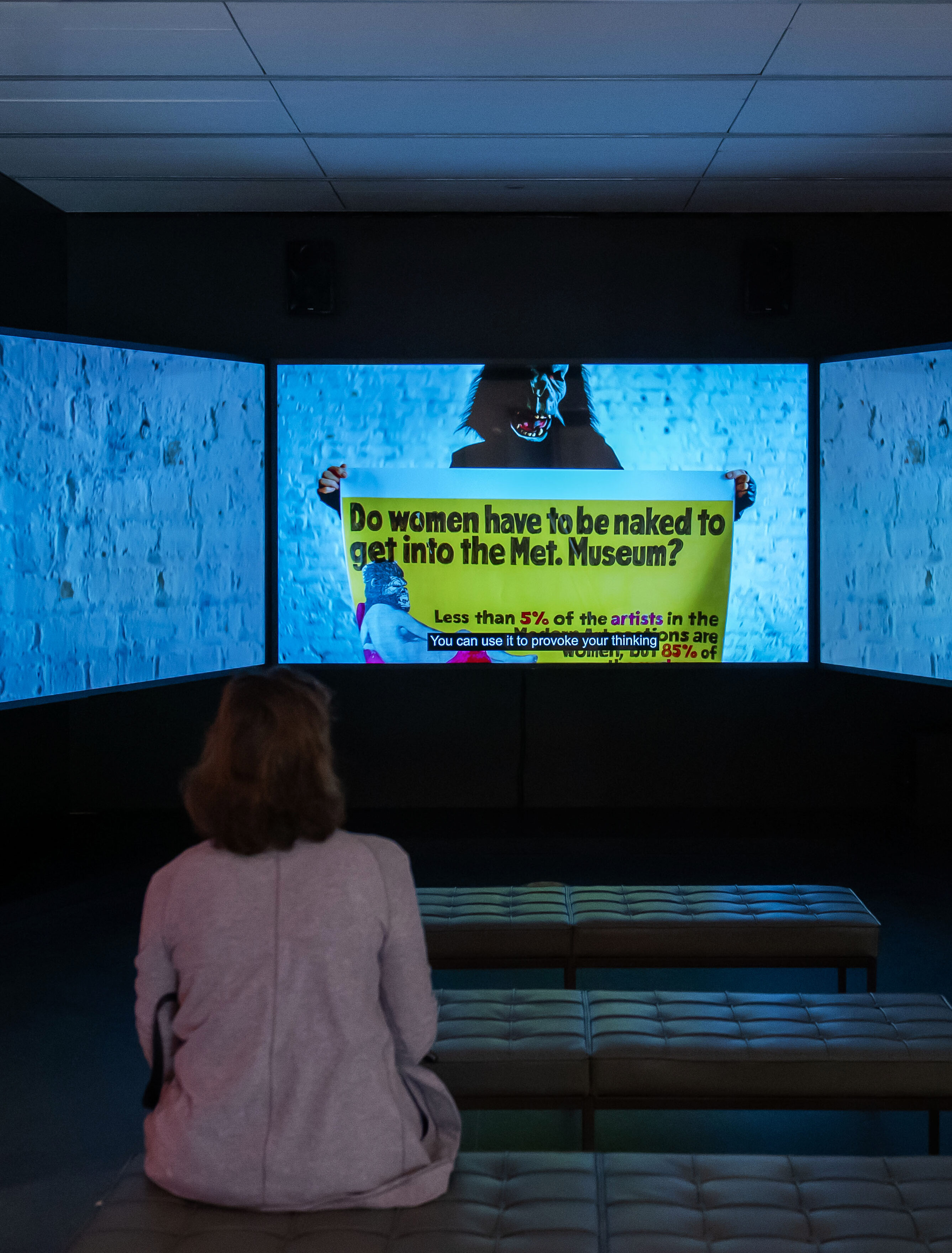 Two women are standing and sitting in a dark room in front of three large screens. A film is playing, showing a person in a gorilla costume holding up a banner. The banner says: 