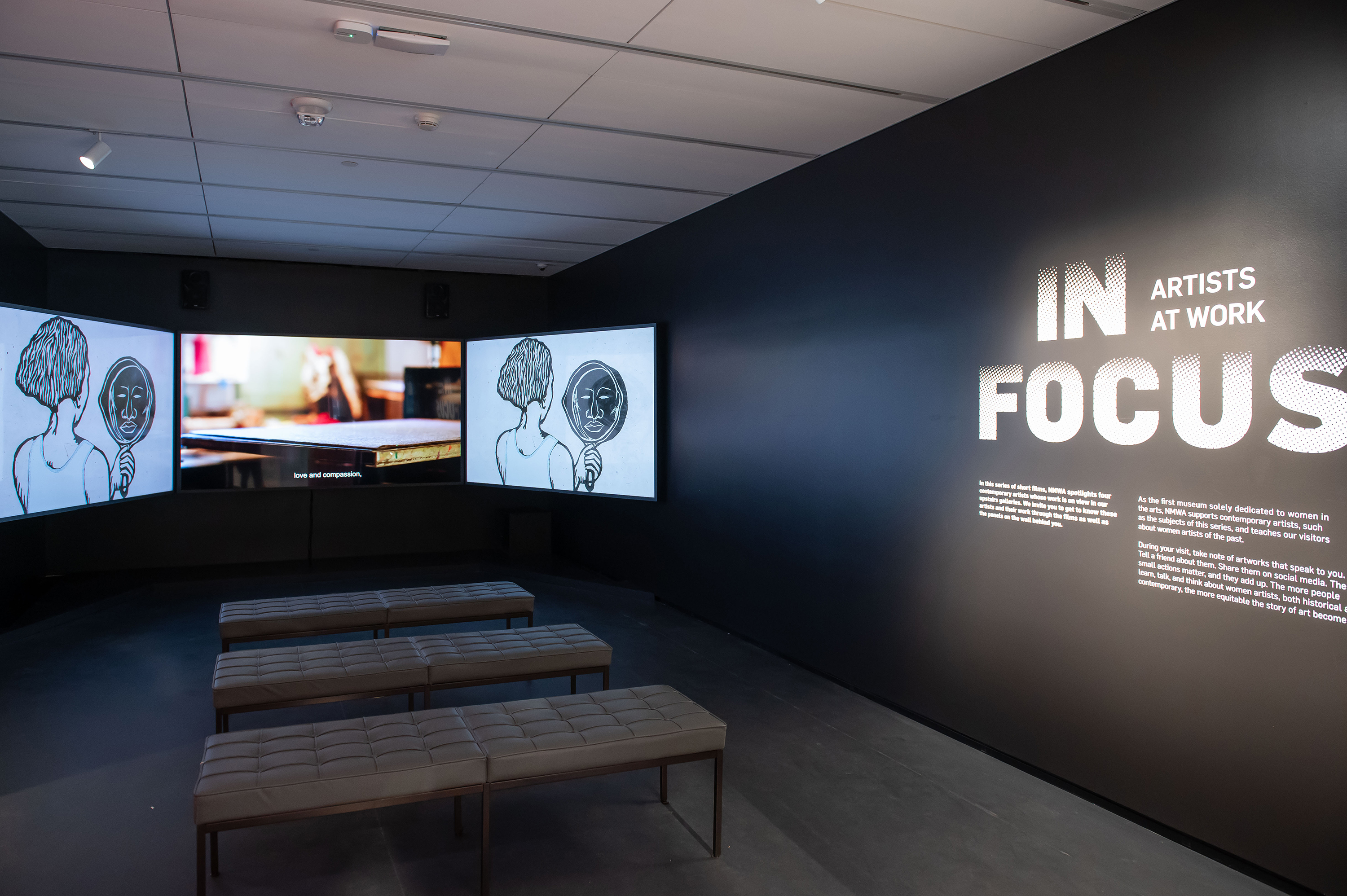 A dark gallery space with three large screens playing a video. Three tufted benches are in front of the screens. On the right wall, it says "In Focus: Artists at Work."