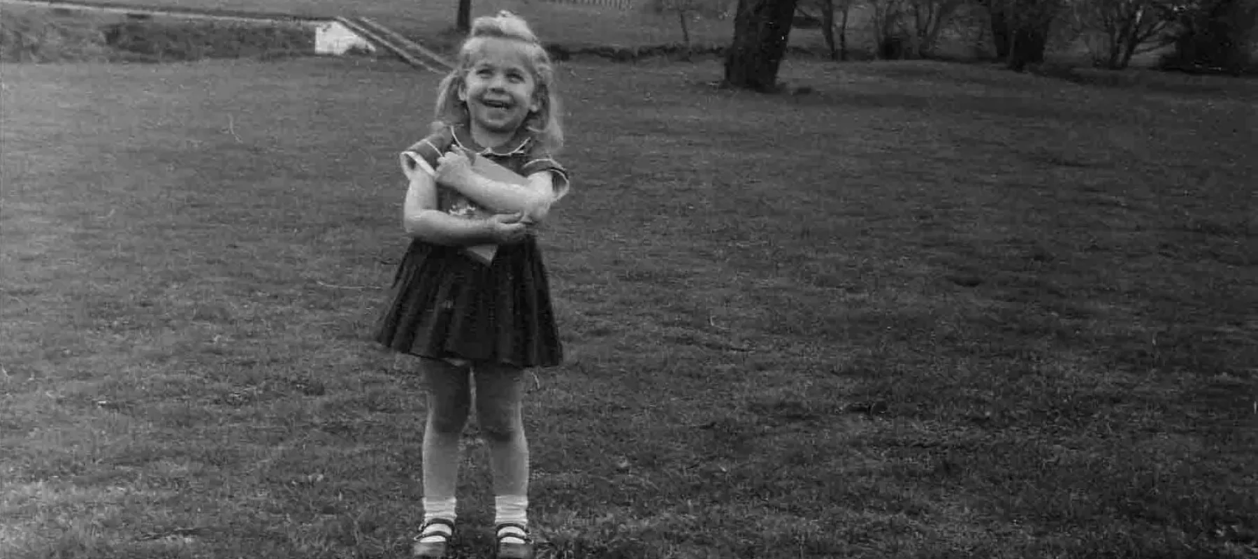 Black and white photograph of the artist as a young girl outdoors hugging an 'lift the flap' book by H. A. Rey.