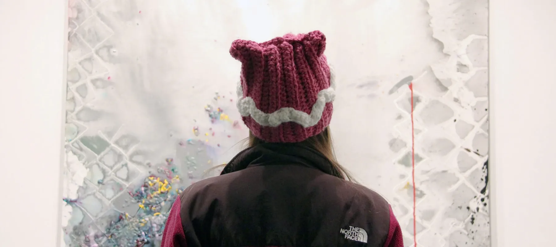 A person in a knitted pink hat in the shape of cat ears is photographed looking at a painting from behind. A mixed media piece featuring colorful, vibrant speckles of color resembling flower buds sprinkled on a gray canvas. They gray canvas has a watercolor texture, and a fence is outlined. The notion of a prison cell comes to mind, as well as hope in the form of color and flowers.