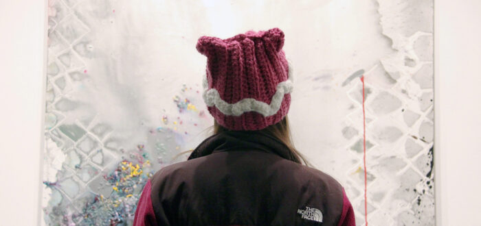 A person in a knitted pink hat in the shape of cat ears is photographed looking at a painting from behind. A mixed media piece featuring colorful, vibrant speckles of color resembling flower buds sprinkled on a gray canvas. They gray canvas has a watercolor texture, and a fence is outlined. The notion of a prison cell comes to mind, as well as hope in the form of color and flowers.