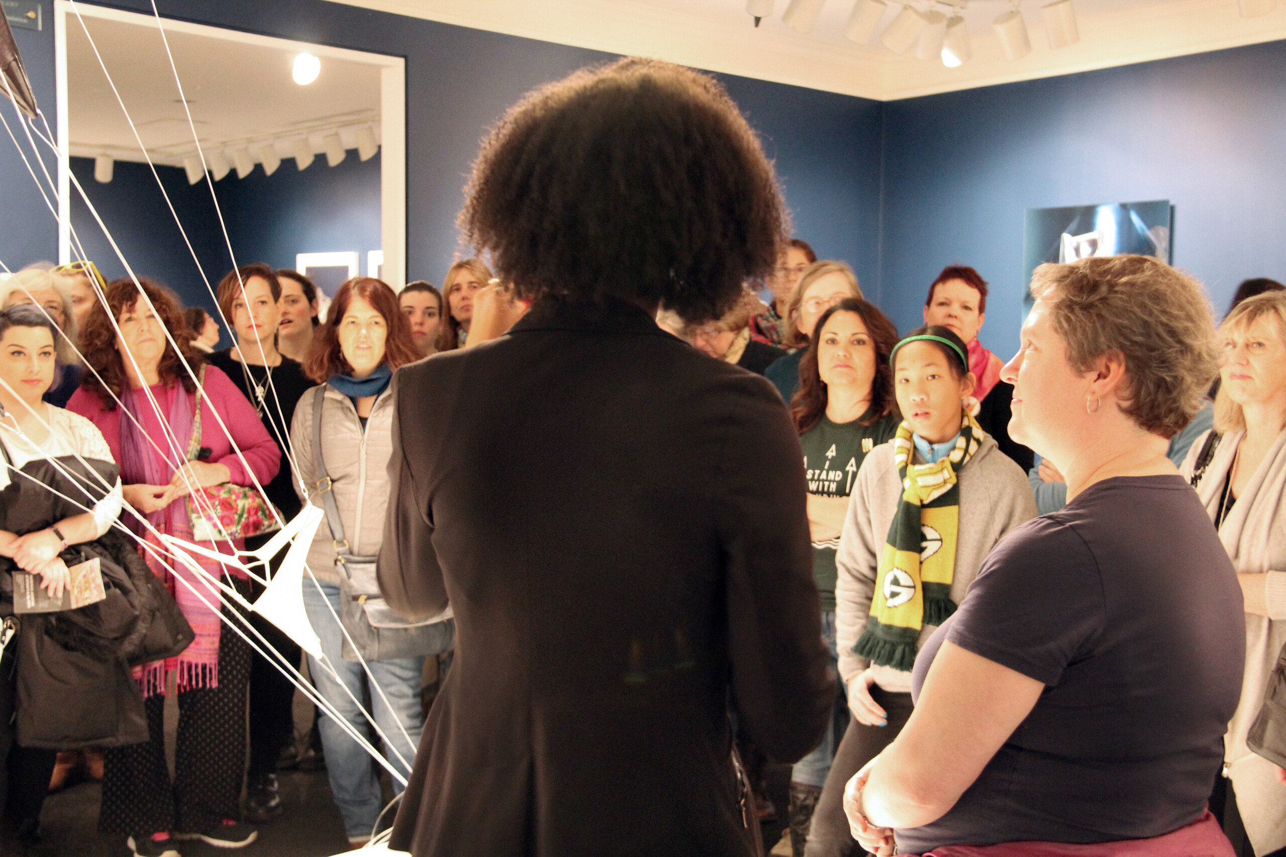 NMWA educator with dark curly hair and a black blazer stands in the foreground with her back to camera as she speaks to a large crowd of visitors about a sculpture composed of hanging strings and fabric in the galleries.