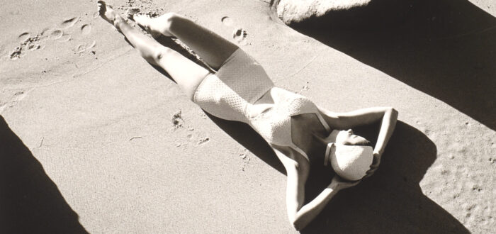 A black and white photograph of a light-skinned woman wearing a one-piece white bathing suit and white swim cap. She is lying down on sand with her hands behind her head. There are footprints in the sand below her, and the image is framed by large rocks.