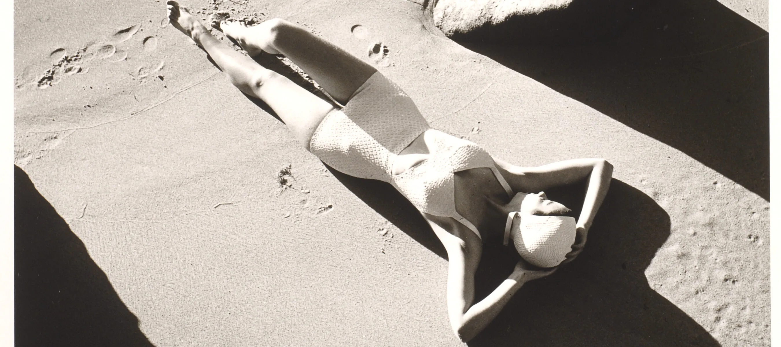 A black and white photograph of a light-skinned woman wearing a one-piece white bathing suit and white swim cap. She is lying down on sand with her hands behind her head. There are footprints in the sand below her, and the image is framed by large rocks.