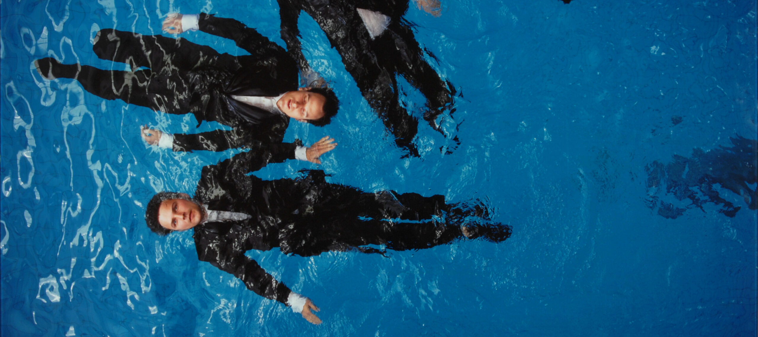 A photograph of four light-skinned figures with short brown hair, wearing black suits with white shirts, floating on their backs in a deep blue pool. The viewer looks down on the figures from above. One figure has their eyes closed, the rest have their eyes open.