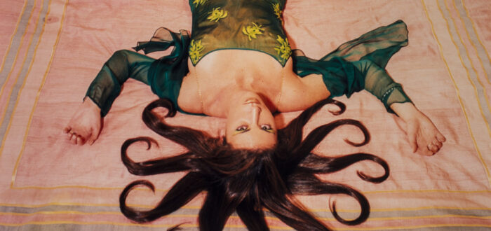 A slim, light-skinned woman wearing a long, emerald-green dress gazes up at the viewer from the foot of the bed, her bare feet near the headboard. Brown hair radiates from her head in 10 curled, snake-like segments. Her arms, bent at the elbows, extend from her body to form a W.