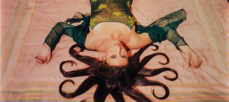 A slim, light-skinned woman wearing a long, emerald-green dress gazes up at the viewer from the foot of the bed, her bare feet near the headboard. Brown hair radiates from her head in 10 curled, snake-like segments. Her arms, bent at the elbows, extend from her body to form a W.