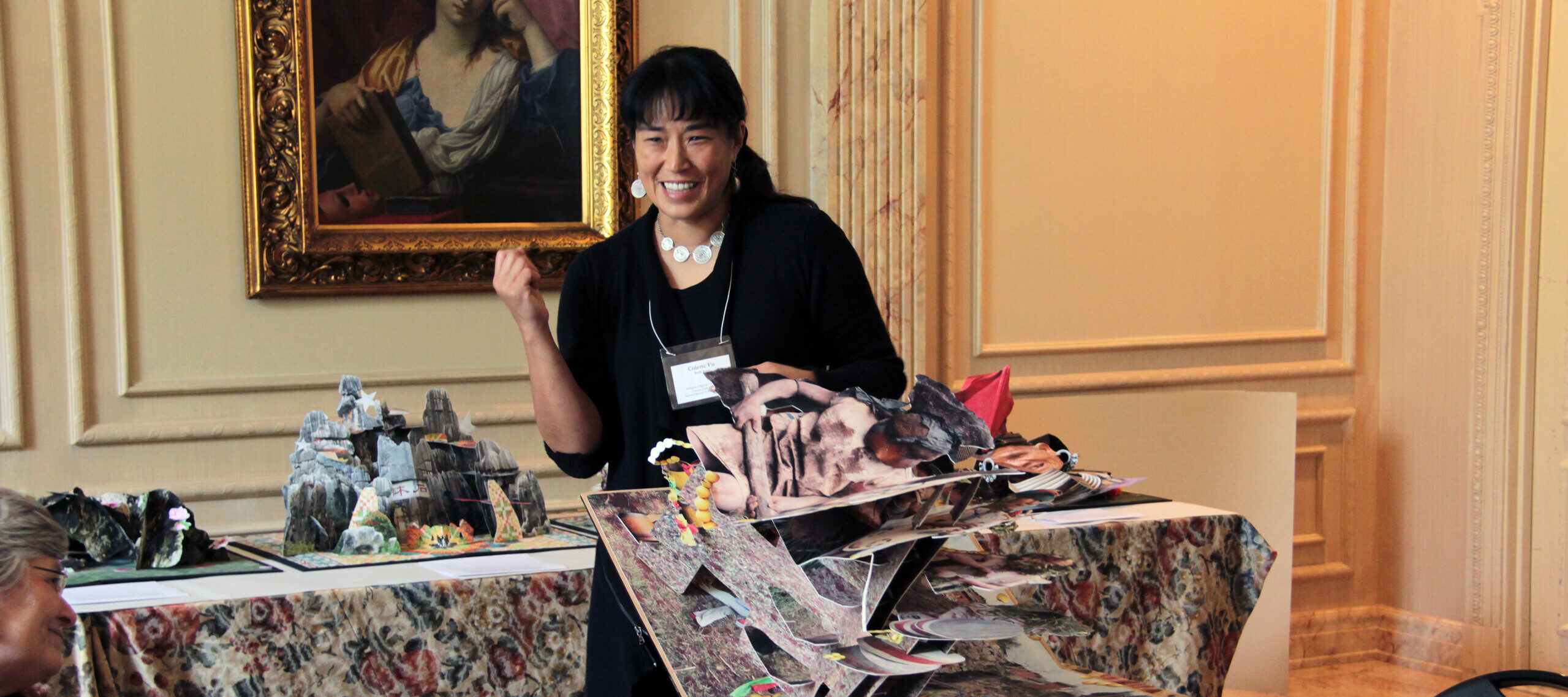 A woman with medium skin tone and dark hair wearing a black dress holds a large pop-up artist book, which unfolds in her hand and onto a table.